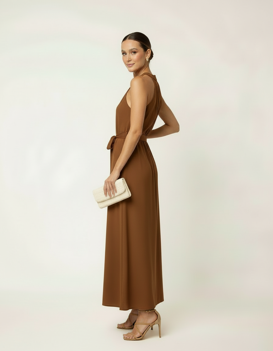 Mulher de lado com vestido halter longo marrom com cinto laço, segurando bolsa clutch branca e sandália bege de salto