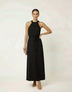 Mulher de frente com vestido halter longo preto com cinto laço e bolsa clutch dourada