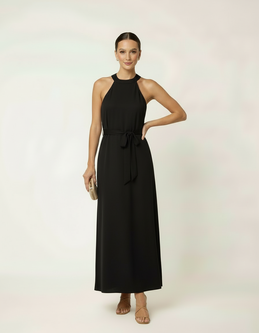 Mulher de frente com vestido halter longo preto com cinto laço e bolsa clutch dourada
