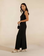 Mulher de lado com vestido longo preto com fenda lateral, bolsa preta e sandália branca de salto